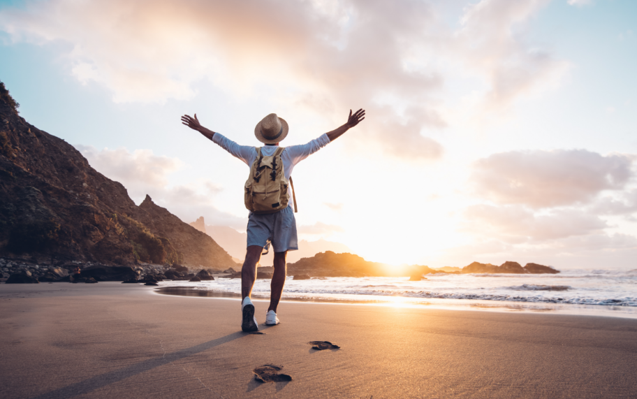 Man walking towards ocean with backpack and hat, arms outstretched